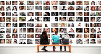 Three people sit on a bench in front of a wall that shows faces of people of different ethnicities, backgrounds and skin colors.